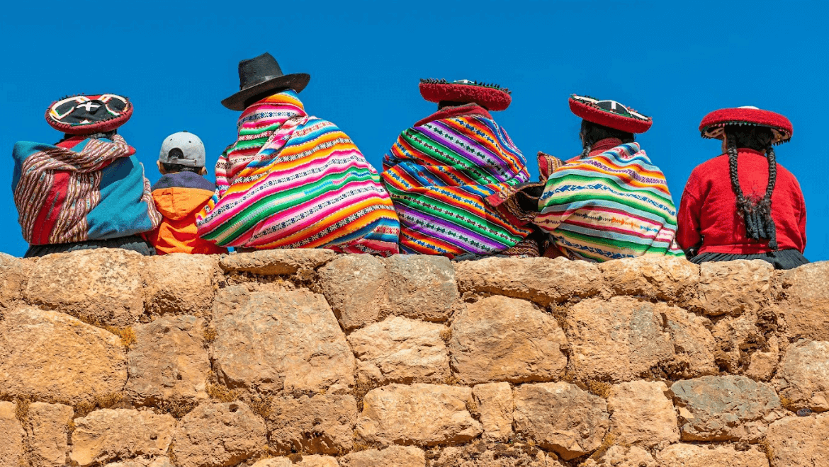 row of people in colorful Peruvian clothes sit on a stone wall facing a bright blue sky in front of them