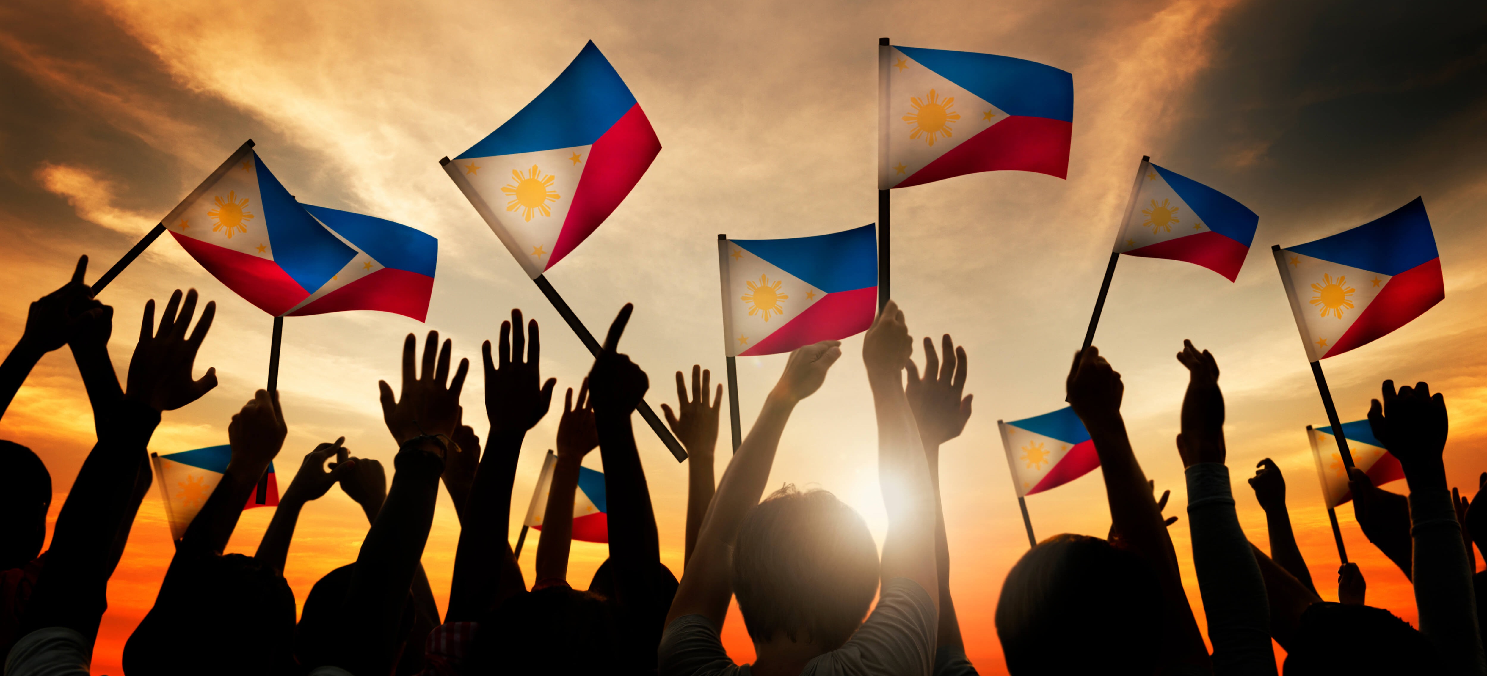 A crowd's silhouetted hands hold up Phillipines flags against a golden sunset